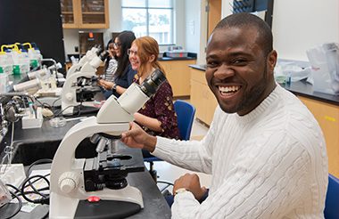Students smiling in a biology lab classroom as they sit in front of microscopes.